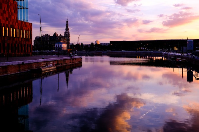 Wolken die reflecteren in het water van een dok met een modern museum en oud havengebouw ernaast.