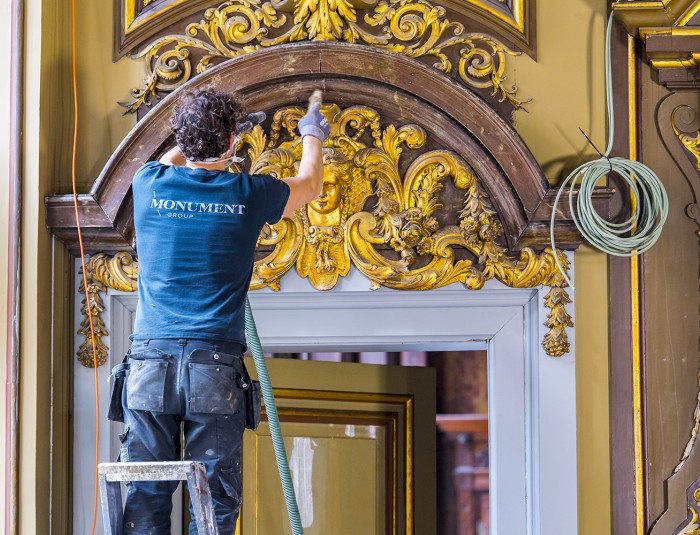 Een restaurator werkt aan ornamenten in bladgoud boven een deur van de collegezaal.